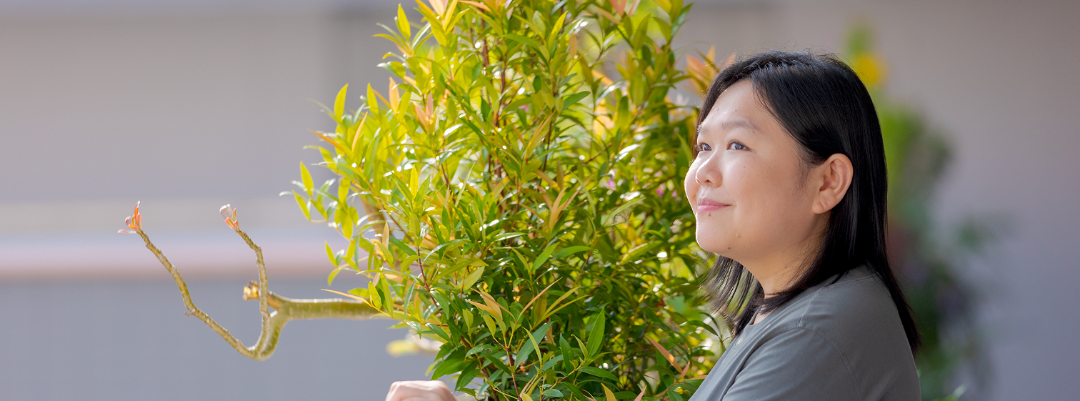 An Asian woman named Andrea with shoulder-length hair and wearing a slate shirt looking off to the left. She is standing in front of a bright green shrub