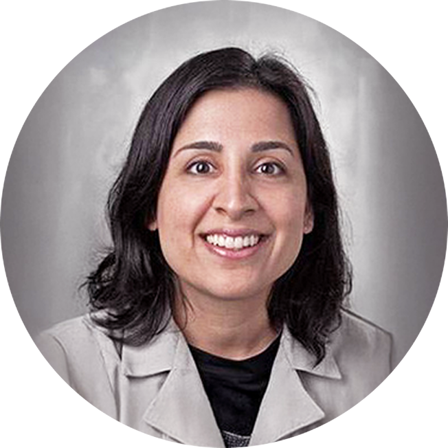 Dr. Amina Ahmed. She has shuolder-length, dark brown hair and is smiling and looking at the viewer. She is wearing a white coat.