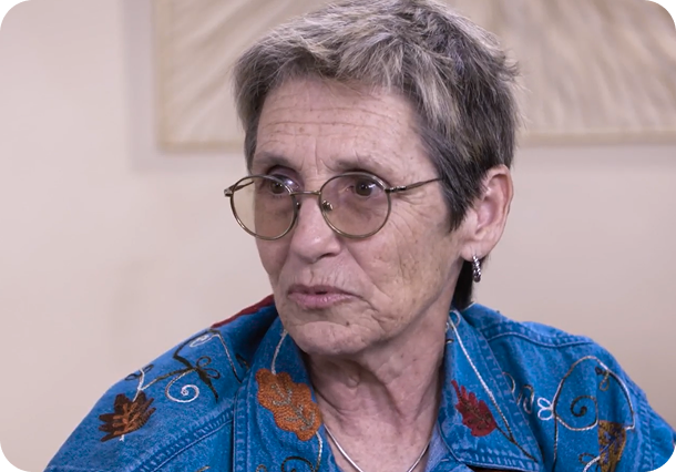 A link to a video. Esther, a myelofibrosis care parther. She is an older white woman with short gray hair and rounded glasses, wearing a denim shirt with floral embroidery. She is speaking with someone to her left.