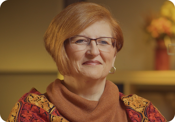 A link to a podcast. An older woman with glasses and short red hair, wearing a scarf and a brightly patterned orange and red top.