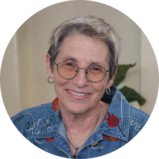 Esther, a myelofibrosis care parther. She is an older woman with short gray hair and rounded glasses, wearing a denim shirt with floral embroidery. She is smiling at the viewer.