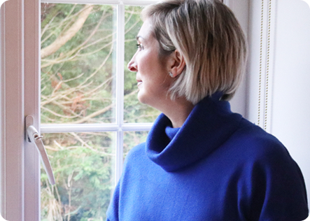 Lou, a woman with short blonde hair facing left, looking thoughtfully out a window.