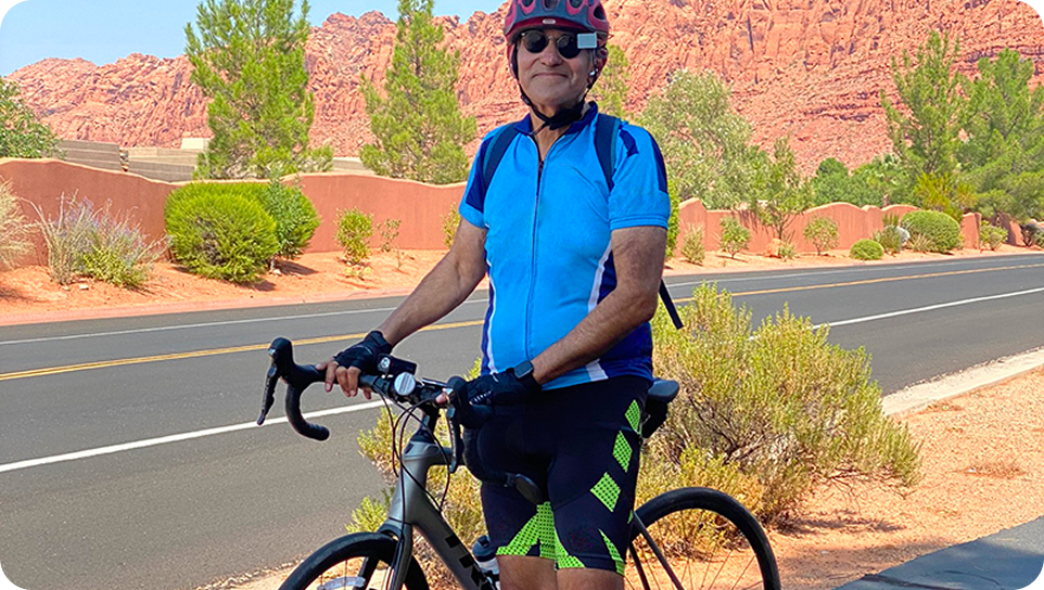 A white man in a blue, black, and neon green biking outfit, standing next to his bike. He is standing in front of a road in bright sunlight, with a desert-like background.