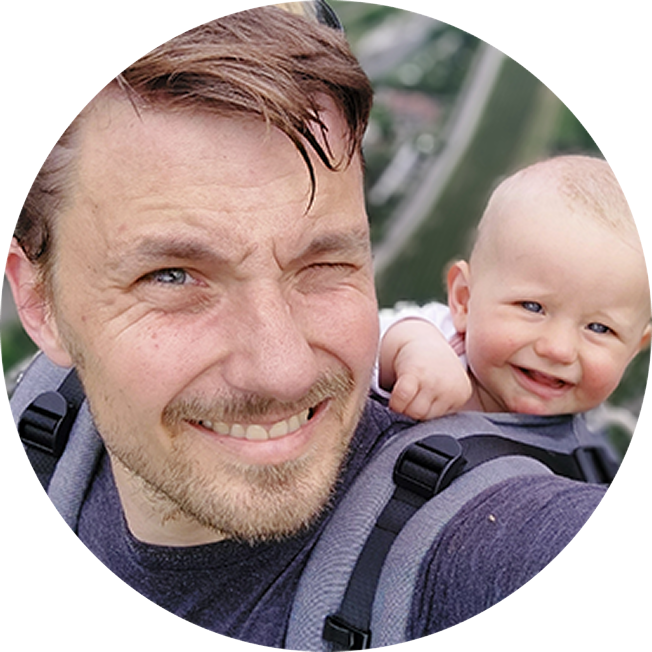 Thomas, a person living with myelofibrosis. He is smiling aat the camera with short brown hair and a short beard, and he is holding his toddler in a backpack. His child is smiling at the camera.