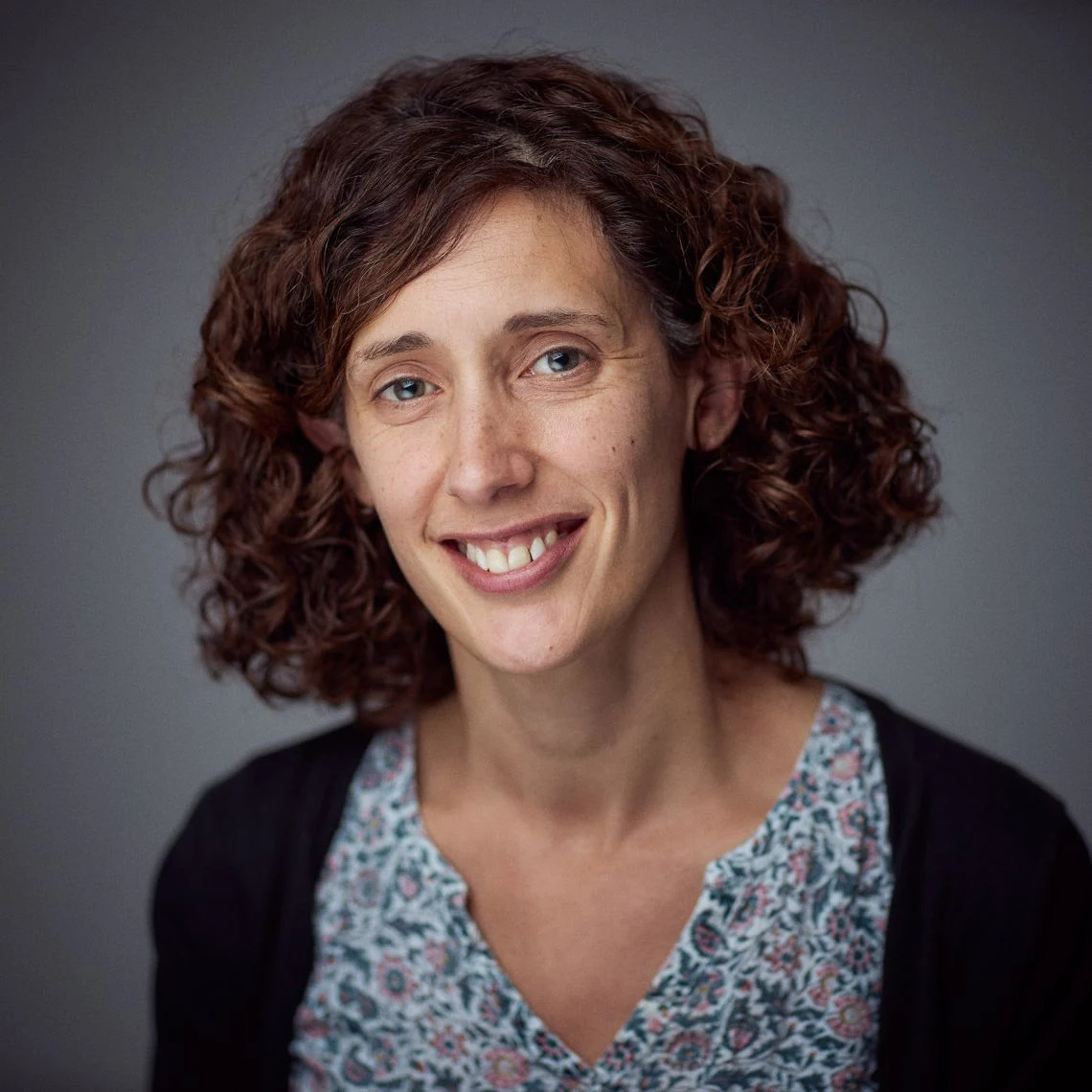 Sarah Gooding, MD, PhD. She is smiling at the viewer and has wavy brown hair. She is wearing a black cardigan over a floral blue top.
