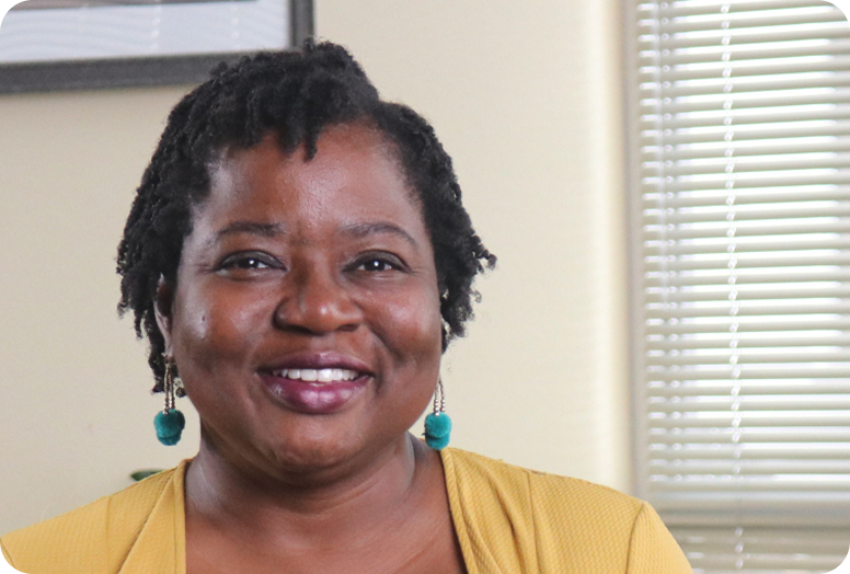 A link to a video. A smiling Black woman named Vernelia with green earrings wearing a yellow cardigan over her black top.