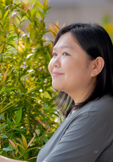 An Asian woman named Andrea with shoulder-length hair and wearing a slate shirt looking off to the left. She is standing in front of a bright green shrub
