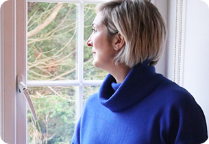Lou, a woman with short blonde hair facing left, looking thoughtfully out a window.
